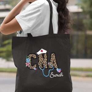 May include: Black tote bag with a leopard print design featuring the letters "CNA" with a stethoscope and a nurse's hat. The name "Sarah" is written below the design.
