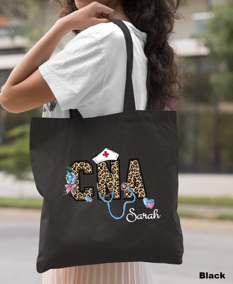 May include: Black tote bag with a leopard print design featuring the letters "CNA" with a stethoscope and a nurse's hat. The name "Sarah" is written below the design.
