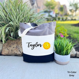 K&ouml;nnte beinhalten: Eine graue, wei&szlig;e und schwarze Canvas-Tragetasche mit dem Namen "Taylor" und einem gelben Pickleball, der auf der Vorderseite aufgedruckt ist. Die Tasche hat einen schwarzen Griff.