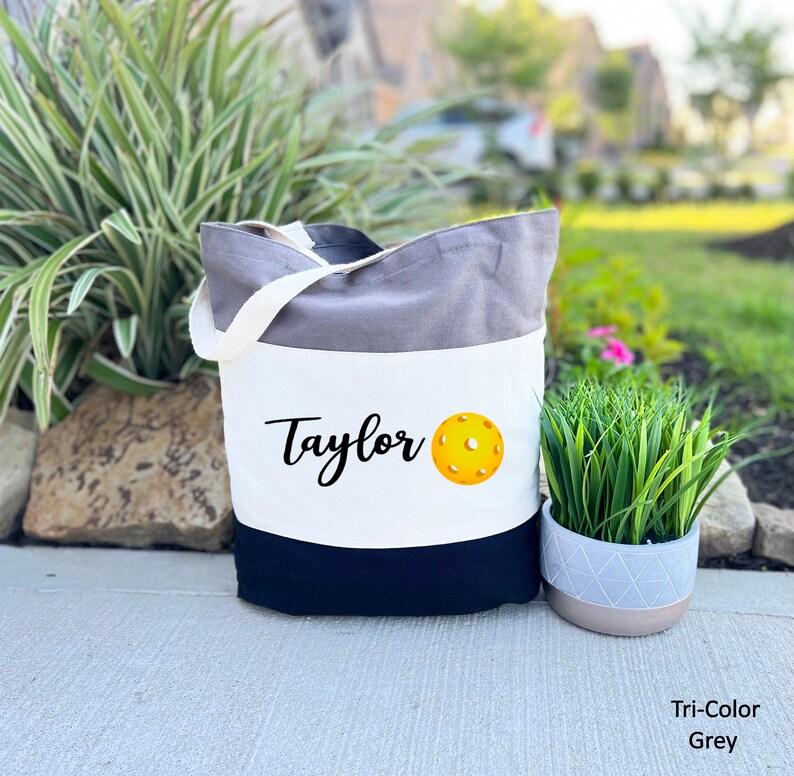 K&ouml;nnte beinhalten: Eine graue, wei&szlig;e und schwarze Canvas-Tragetasche mit dem Namen "Taylor" und einem gelben Pickleball, der auf der Vorderseite aufgedruckt ist. Die Tasche hat einen schwarzen Griff.