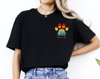 May include: Black t-shirt featuring a colorful paw print design with a stethoscope and the name "Sarah". The paw print incorporates shades of red, orange, yellow, and green, creating a retro aesthetic. The shirt is a casual wear item.