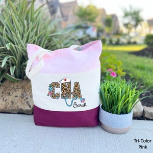 May include: A tri-color tote bag with a pink, white, and burgundy design. The bag features a leopard print "CNA" with a stethoscope and floral accents. The name "Sarah" is printed below the design.