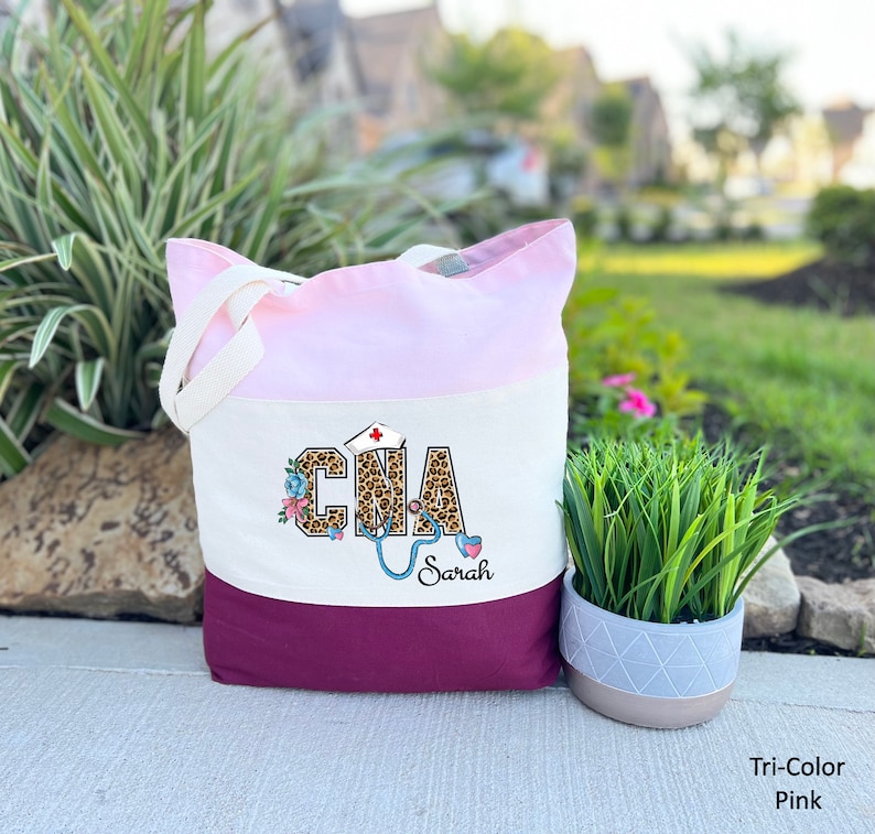 May include: A tri-color tote bag with a pink, white, and burgundy design. The bag features a leopard print "CNA" with a stethoscope and floral accents. The name "Sarah" is printed below the design.