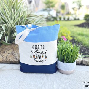 Pode incluir: Uma bolsa tote tricolor com um design azul, branco e azul marinho. A bolsa tem uma se&ccedil;&atilde;o de lona branca com o texto "Easily Distracted By Plants" e um gr&aacute;fico de plantas penduradas.