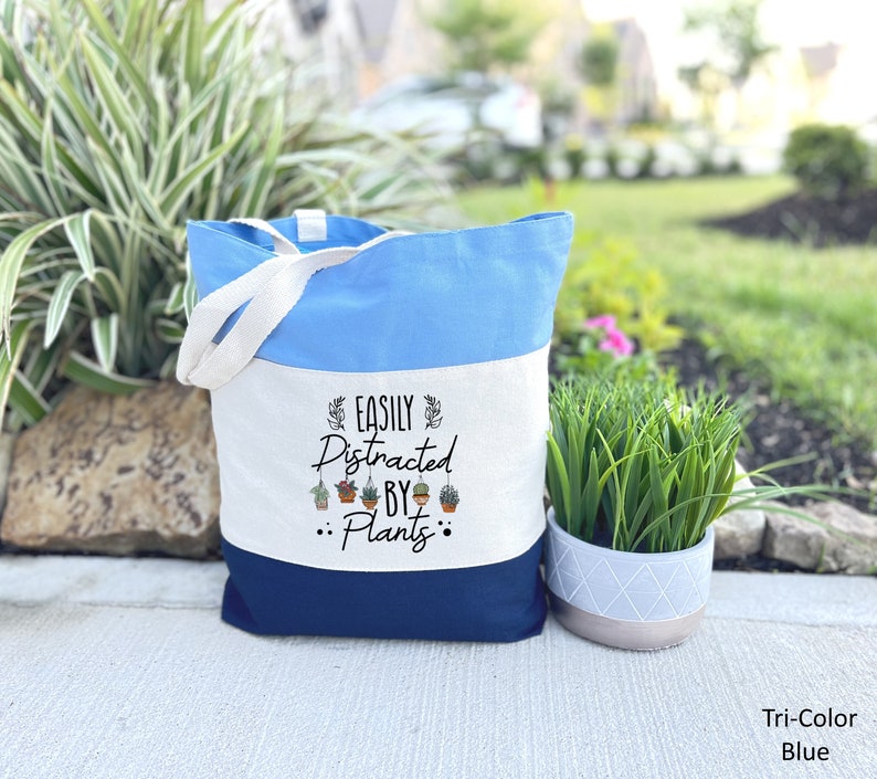 Pode incluir: Uma bolsa tote tricolor com um design azul, branco e azul marinho. A bolsa tem uma se&ccedil;&atilde;o de lona branca com o texto "Easily Distracted By Plants" e um gr&aacute;fico de plantas penduradas.