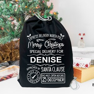 May include: Black Christmas gift bag with white text that reads "Express Delivery North Pole Merry Christmas Special Delivery for Denise Approved by: Santa Clause List 2x Checked Do Not Open Before 25 December".
