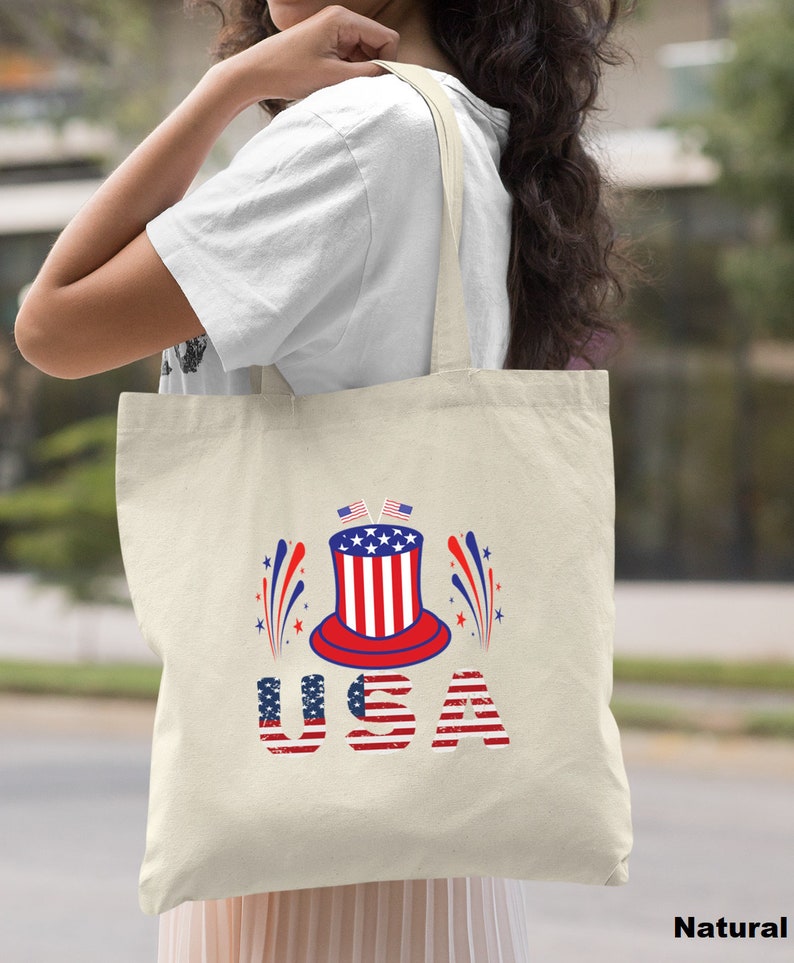May include: A natural canvas tote bag with a red, white, and blue graphic of a top hat with fireworks and the word "USA" in red, white, and blue.