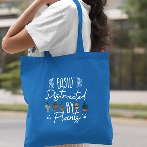 Pode incluir: Uma bolsa tote azul royal com texto branco que diz "Easily Distracted By Plants". O texto &eacute; cercado por pequenos desenhos de plantas em vasos.