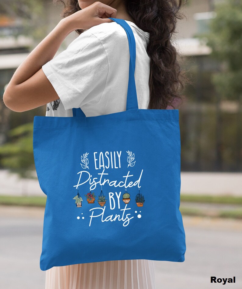 Pode incluir: Uma bolsa tote azul royal com texto branco que diz "Easily Distracted By Plants". O texto &eacute; cercado por pequenos desenhos de plantas em vasos.