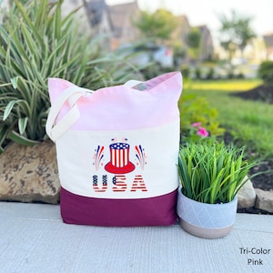 May include: A pink, white, and burgundy canvas tote bag with a red, white, and blue graphic of a top hat with fireworks and the word "USA" printed on it.