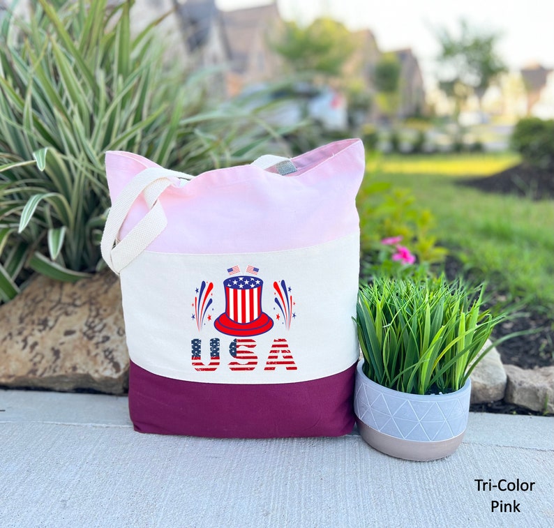 May include: A pink, white, and burgundy canvas tote bag with a red, white, and blue graphic of a top hat with fireworks and the word "USA" printed on it.