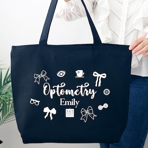May include: A navy blue tote bag with white graphics and the word "Optometry" in script. The bag features eye-related icons and the name "Emily". The bag has two black handles.