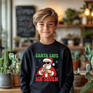 May include: Black sweatshirt with a graphic of Santa Claus wearing sunglasses and making a hand gesture. The text "SANTA SAYS" is above the Santa graphic, and "SIX SEVEN" is below it. The sweatshirt is worn by a young person.