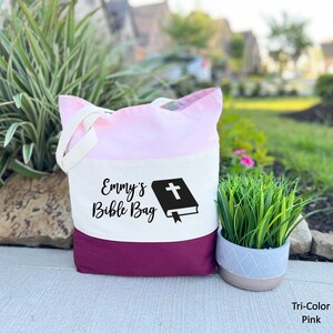 May include: A tri-color tote bag with a pink, white, and burgundy design. The bag has a white cross on a black bible graphic and the text "Emmy's Bible Bag".
