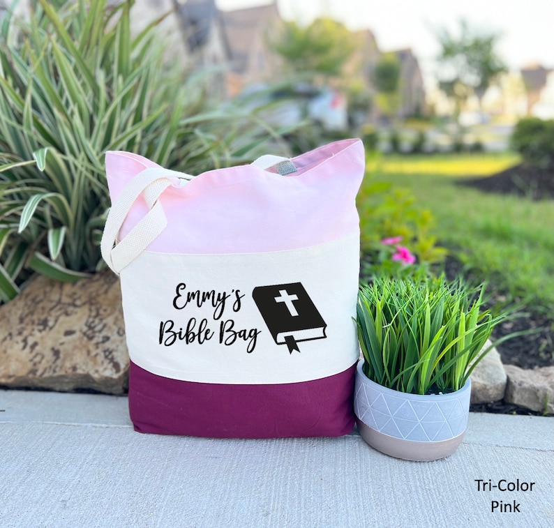 May include: A tri-color tote bag with a pink, white, and burgundy design. The bag has a white cross on a black bible graphic and the text "Emmy's Bible Bag".
