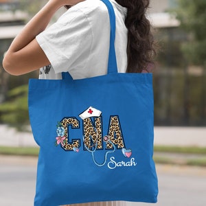 May include: A royal blue tote bag with a leopard print design that says "CNA" with a stethoscope and a nurse's hat. The name "Sarah" is written below the design.