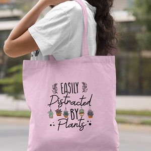 Pode incluir: Uma bolsa tote rosa claro com o texto "EASILY DISTRACTED BY PLANTS" e um gr&aacute;fico de plantas em vasos pendurados.