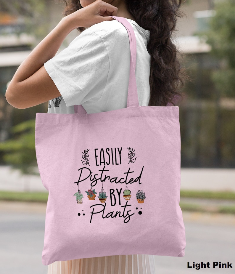 Pode incluir: Uma bolsa tote rosa claro com o texto "EASILY DISTRACTED BY PLANTS" e um gr&aacute;fico de plantas em vasos pendurados.