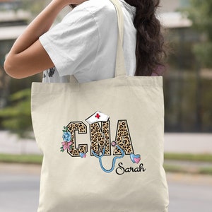 May include: A natural canvas tote bag with a leopard print design that says "CNA" with a stethoscope and a red cross on a white hat. The name "Sarah" is written below the design.