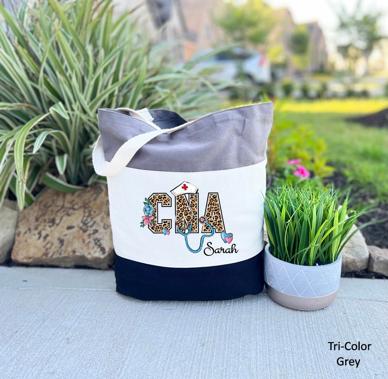 May include: A tri-color tote bag with a grey, white, and black design. The white section features a leopard print "CNA" with a stethoscope and flowers. The text "Sarah" is below the design.