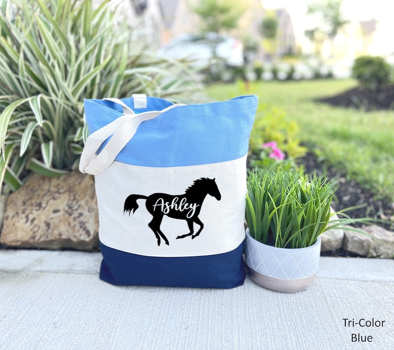May include: A blue, white, and navy blue tote bag with a silhouette of a horse and the name "Ashley" printed on the front.