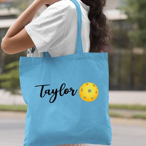 K&ouml;nnte beinhalten: Hellblaue Canvas-Tragetasche mit dem Namen "Taylor" und einer gelben Pickleball-Grafik.