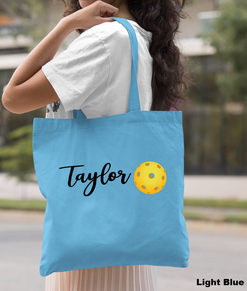 K&ouml;nnte beinhalten: Hellblaue Canvas-Tragetasche mit dem Namen "Taylor" und einer gelben Pickleball-Grafik.