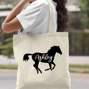 May include: A natural canvas tote bag with a black silhouette of a horse running and the name "Ashley" written on the horse's side.
