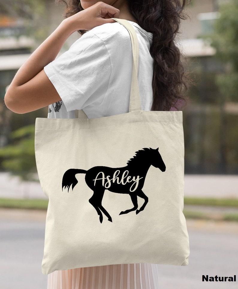 May include: A natural canvas tote bag with a black silhouette of a horse running and the name "Ashley" written on the horse's side.