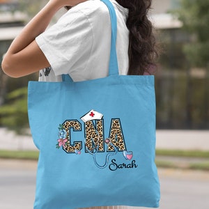 May include: Light blue tote bag with a leopard print "CNA" design, a stethoscope, a red cross, and flowers. The name "Sarah" is written below the design.