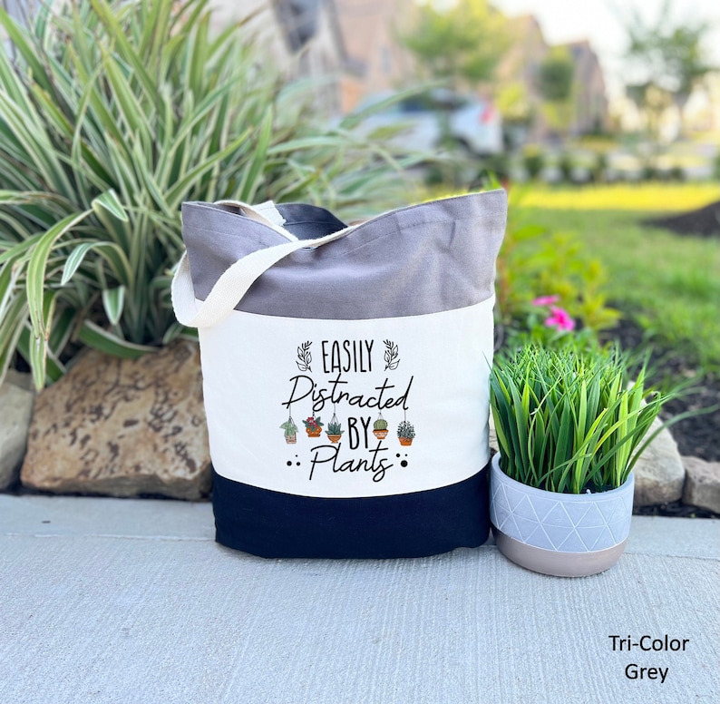 Pode incluir: Uma bolsa de lona branca com al&ccedil;as pretas e fundo preto. A bolsa tem uma faixa branca com o texto "EASILY DISTRACTED BY PLANTS" e uma fileira de pequenas plantas em vasos. A bolsa est&aacute; sobre uma superf&iacute;cie de concreto cinza.