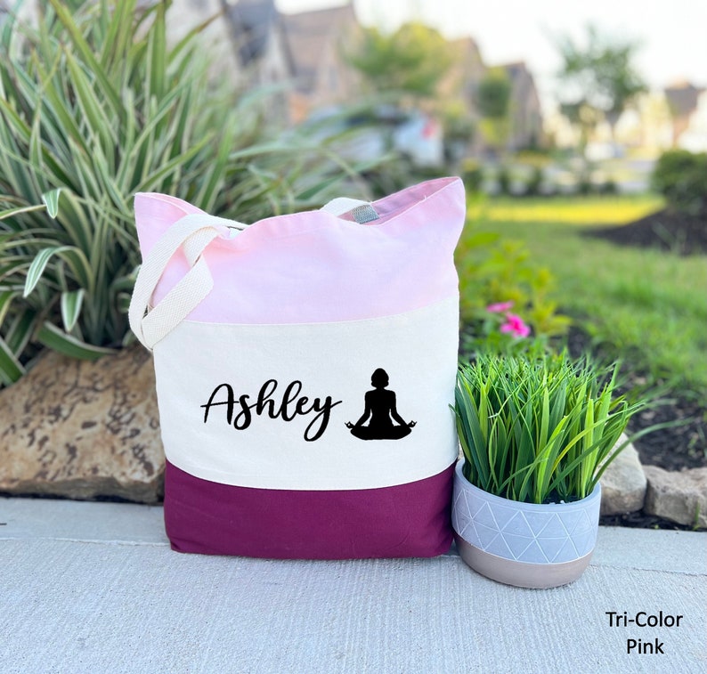Pu&ograve; includere: Una borsa tote tricolore in tessuto rosa, bianco e bordeaux. La borsa ha un pannello bianco con il nome "Ashley" e una silhouette nera di una persona in una posa yoga.