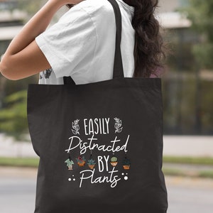 Pode incluir: Sacola de tecido preta com texto branco que diz "Easily Distracted By Plants" com uma fileira de plantas em vaso acima da palavra "Plants".