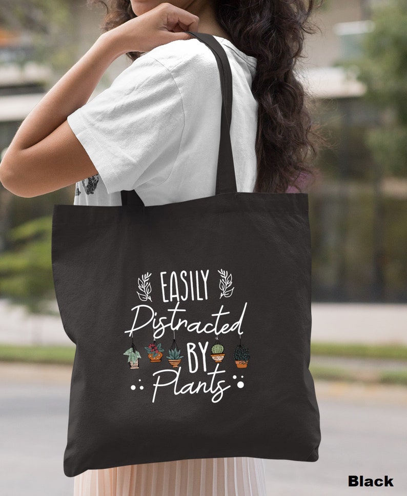 Pode incluir: Sacola de tecido preta com texto branco que diz "Easily Distracted By Plants" com uma fileira de plantas em vaso acima da palavra "Plants".
