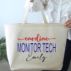May include: A beige canvas tote bag with long handles. The bag features the words "cardiac MONITOR TECH Emily" in red, blue, and black lettering. The bag is held by a person.