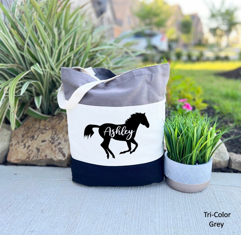 May include: A tri-color tote bag with a grey, white, and black design. The bag features a black silhouette of a horse with the name "Ashley" written on it.