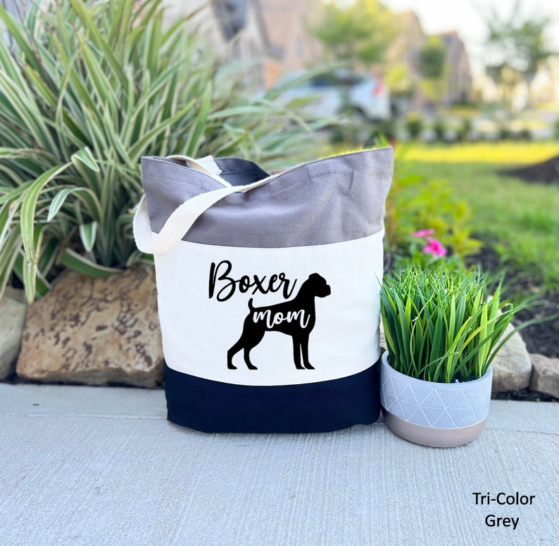 Puede incluir: Una bolsa de mano gris de tres colores con un panel blanco que presenta una silueta negra de un perro boxer y el texto "Boxer mom".
