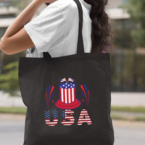 May include: Black tote bag with a red, white, and blue graphic of a top hat with fireworks and the word "USA" in distressed lettering.