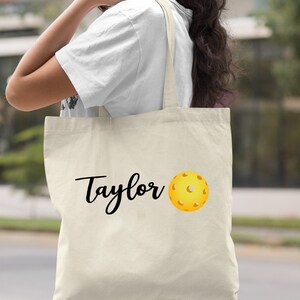 K&ouml;nnte beinhalten: Eine nat&uuml;rliche Canvas-Tragetasche mit schwarzem Text, der "Taylor" sagt, und einer gelben Pickleball-Grafik.