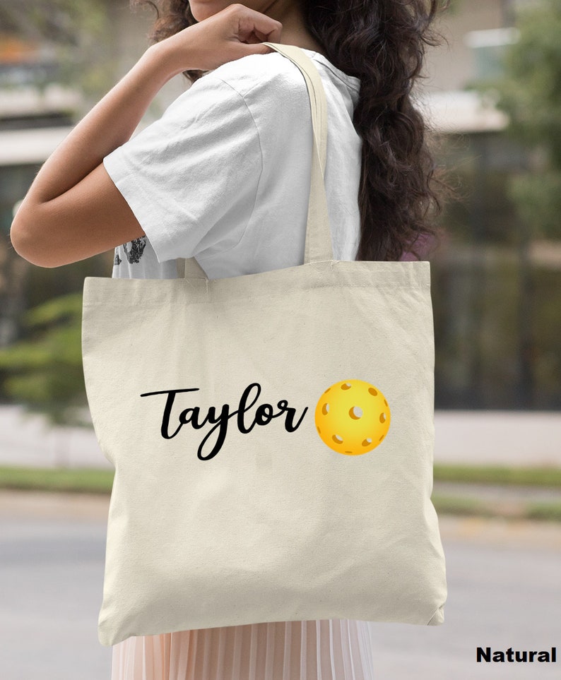 K&ouml;nnte beinhalten: Eine nat&uuml;rliche Canvas-Tragetasche mit schwarzem Text, der "Taylor" sagt, und einer gelben Pickleball-Grafik.