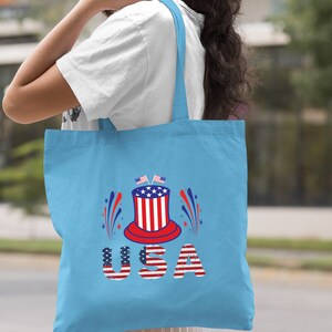 May include: A light blue tote bag with a red, white, and blue graphic of a top hat with fireworks and the word "USA" in distressed lettering.