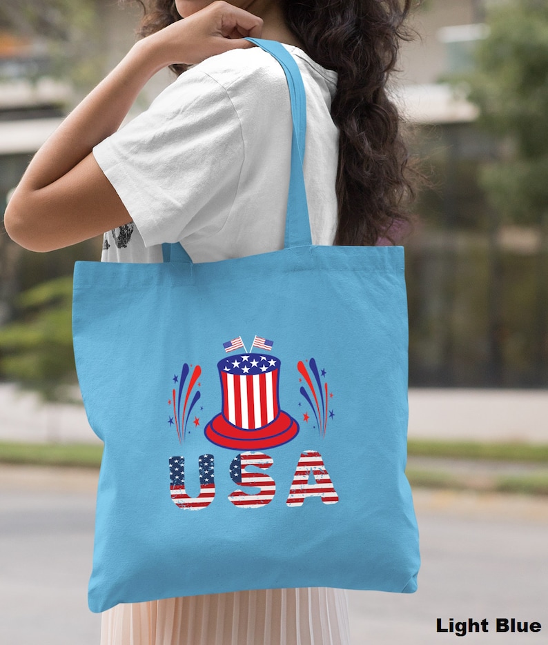 May include: A light blue tote bag with a red, white, and blue graphic of a top hat with fireworks and the word "USA" in distressed lettering.