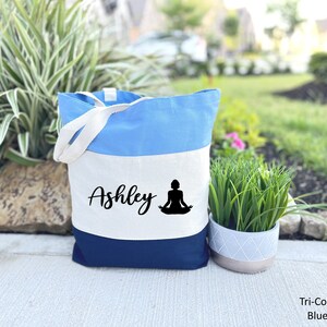 Pu&ograve; includere: Una borsa tote blu, bianca e blu navy con il nome "Ashley" e una silhouette di una posa di yoga stampata sul davanti.