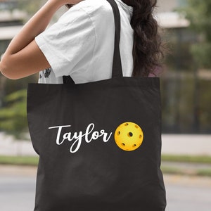 K&ouml;nnte beinhalten: Schwarze Stofftasche mit wei&szlig;em Text "Taylor" und einem gelben Pickleball.