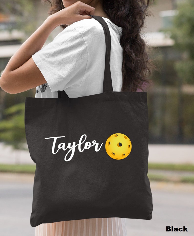 K&ouml;nnte beinhalten: Schwarze Stofftasche mit wei&szlig;em Text "Taylor" und einem gelben Pickleball.
