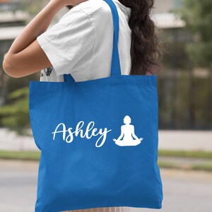 Pu&ograve; includere: Una borsa tote blu reale con manici bianchi e un design bianco di una donna in una posa yoga e il nome "Ashley".