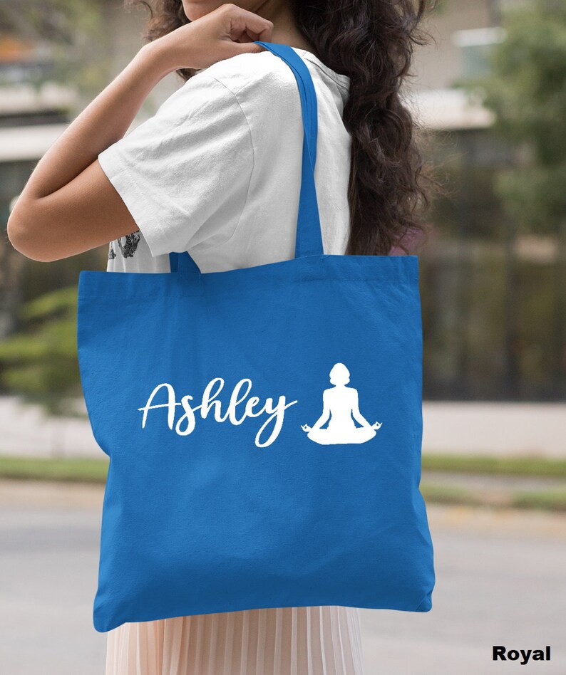 Pu&ograve; includere: Una borsa tote blu reale con manici bianchi e un design bianco di una donna in una posa yoga e il nome "Ashley".