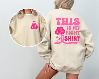 May include: A beige sweatshirt with a pink ribbon and boxing gloves graphic on the back. The text "This is my fight shirt" is printed in pink on the back of the sweatshirt.