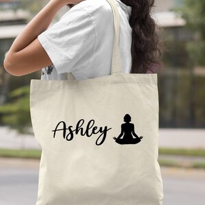 Pu&ograve; includere: Una borsa tote in tela naturale con testo nero e una silhouette di una persona in una posa yoga. Il testo sulla borsa dice "Ashley".