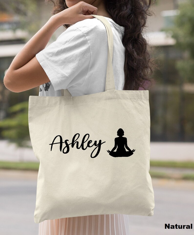 Pu&ograve; includere: Una borsa tote in tela naturale con testo nero e una silhouette di una persona in una posa yoga. Il testo sulla borsa dice "Ashley".